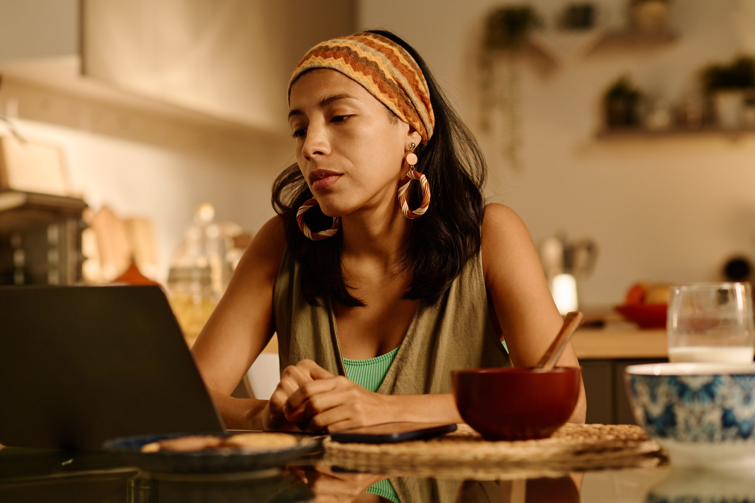 Young stylish businesswoman in homewear looking at laptop screen while sitting by kitchen table after breakfast and working over new project